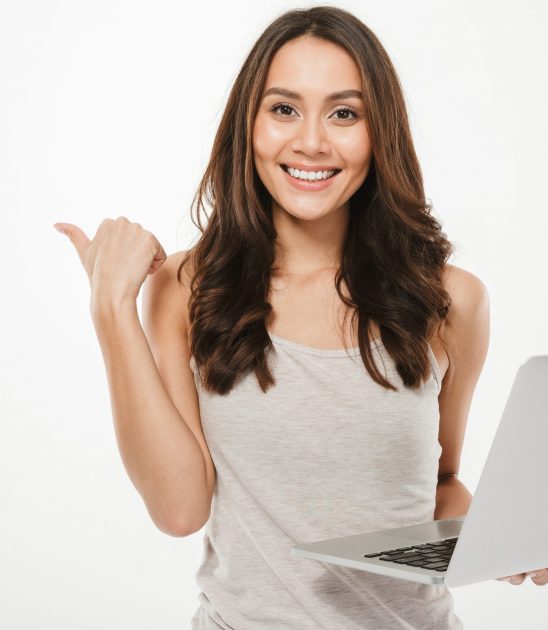 Image of amazing businesswoman holding silver laptop and pointing finger sideways with smile over white wall