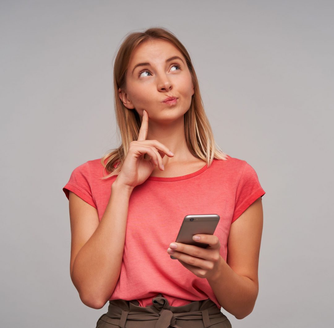 Portrait of cute, wondering girl with blond hair. Wearing pink t-shirt and brown skirt. Holding cellphone and touching her cheek. Watching to the right upper corner at copy space over grey background